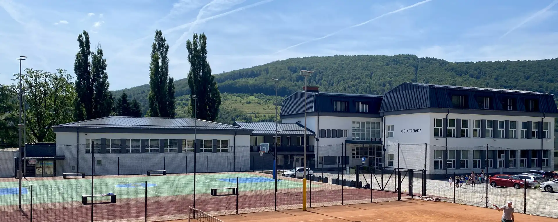 CIK-Trebnje-o-nas Fotografija zavoda CIK Trebnje na lep sončen dan. Pred centrom gruča otrok, v ozadju lep zelen hrib in modro nebo.