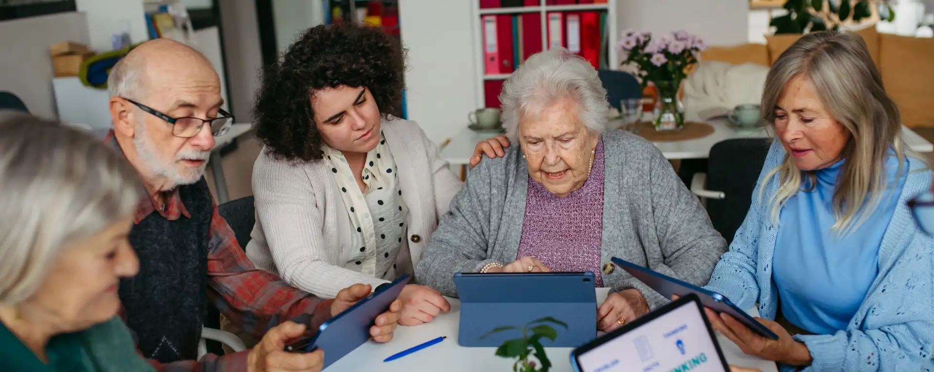 Starejši na delavnici digitalnega usposabljanja. V rokah držijo tablične računalnike, med tem ko jih mentorica uči uporabe.